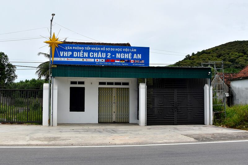 A large blue sign with white lettering naming the building in Vietnamese is posted on top of a small white building with a green roof.