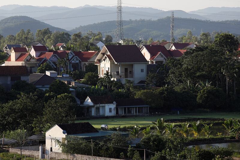 A cluster of large white houses with red roofs tower behind two small, older looking homes amidst lush green trees.