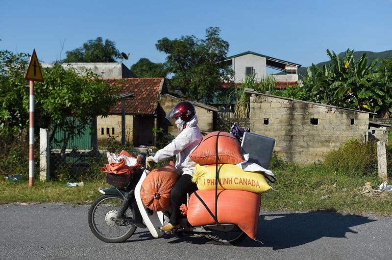 A woman in a white jacket and red helmet rides a motorcycle with large orange and yellow bags strapped to it. One of these bags sits tightly in her lap.